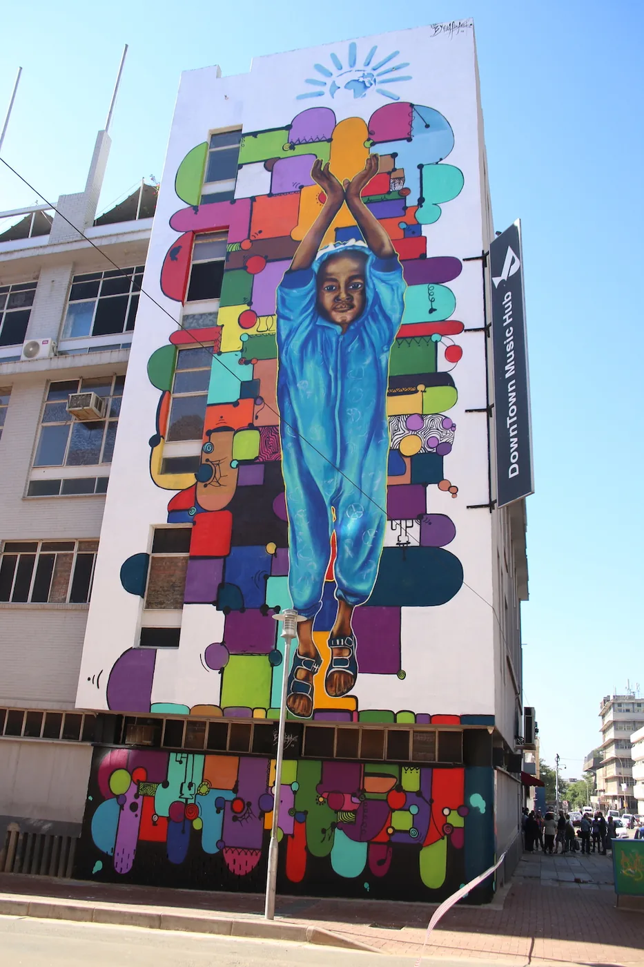 A tall mural on the side of a Johannesburg building shows a figure in blue with raised arms, surrounded by colourful speech bubbles.